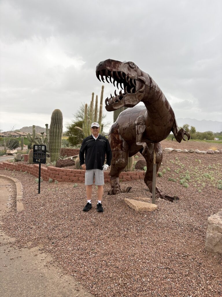 Craig Spiesman posing near a tall dinosaur sculpture with cacti and rocky ground